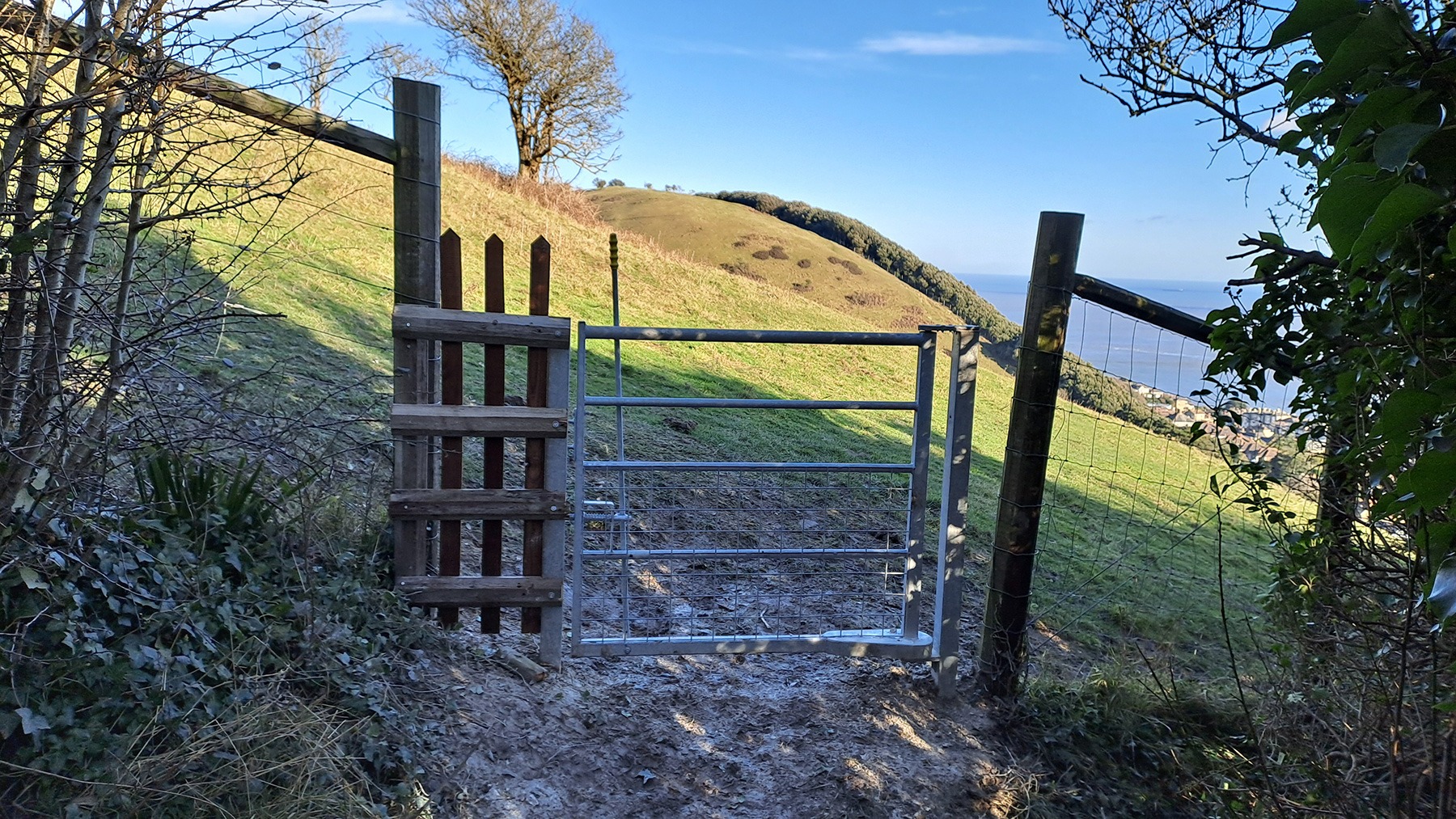 Photo of view through metal gate onto green sloping farmland with blue sky and sea in the background.