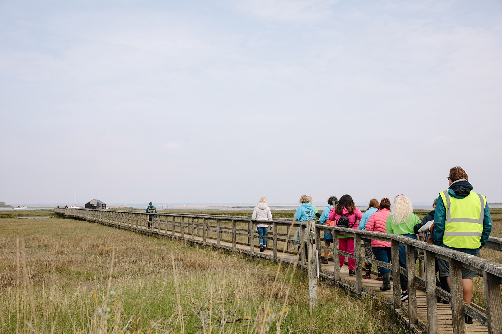 Walkers on a long wooden bridge walkway across a marsh looking towards Newtown Creek
