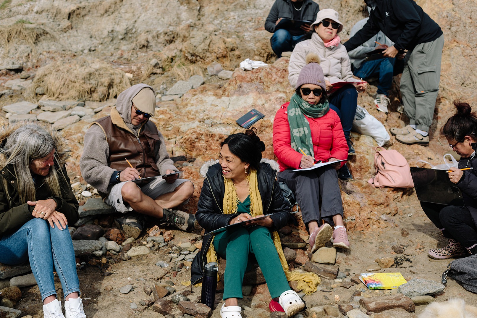 A group sitting on a rocky beach in winter clothes sketching beach views