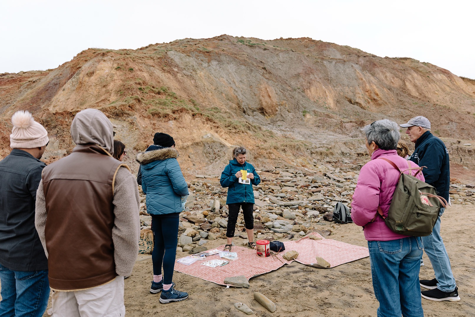 Nature expert holding plant identification card on beach with warm coloured rocks behind and interested group members surrounding her