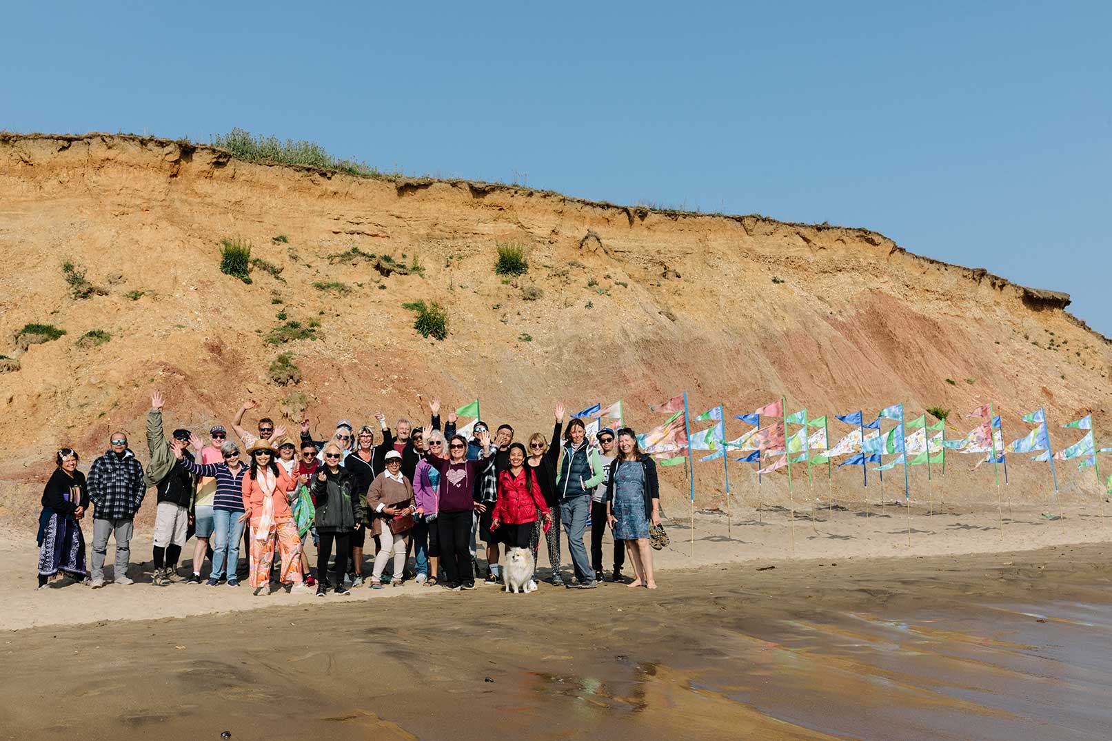 Community group on beach next to colourful pennant flags designed and painted by local people with orange cliff behind and blue sky