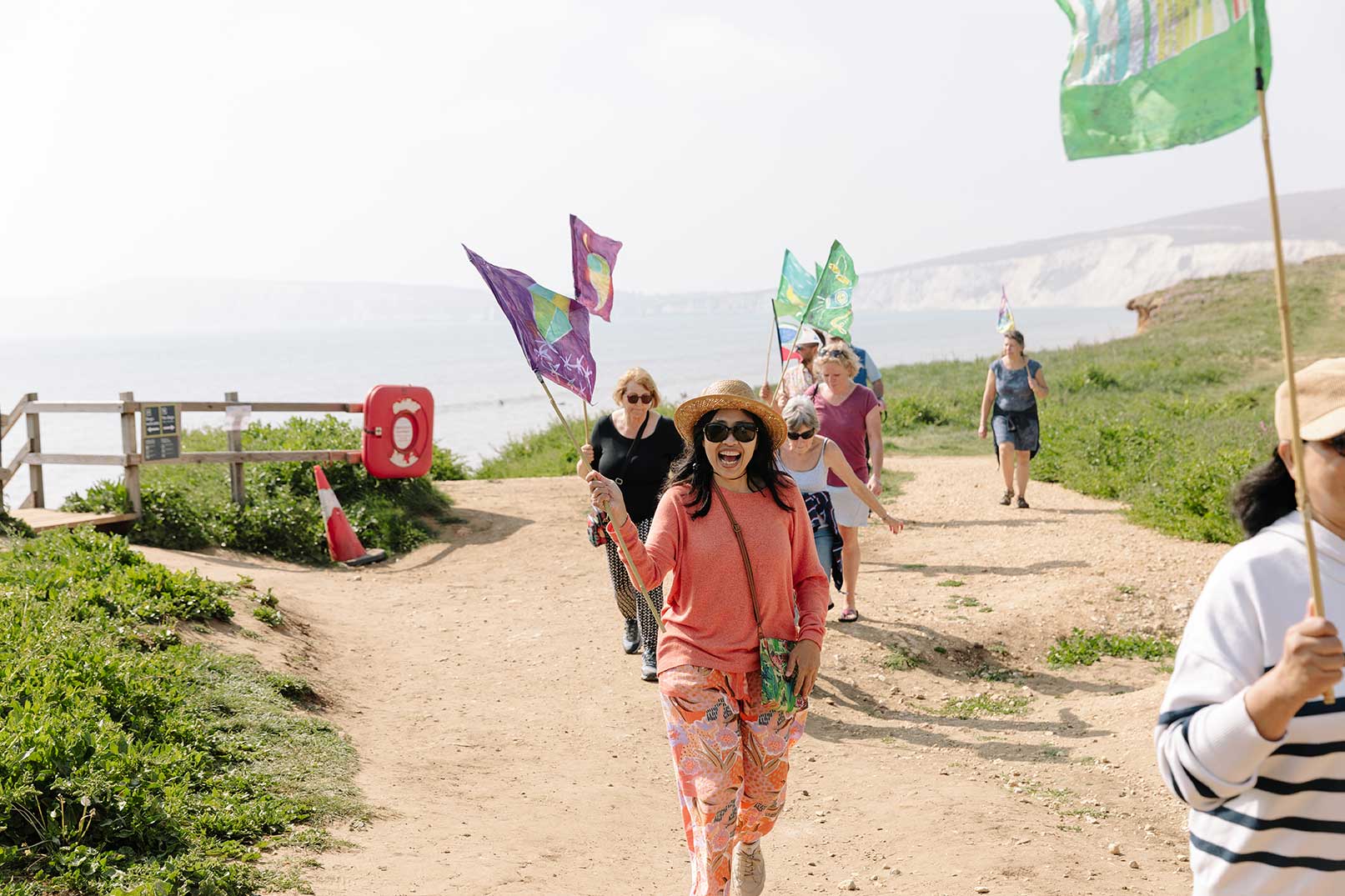 A small group of walks with a happy smiling walker in front of the group waving a hand painted flag along a coastal path with sea and chalk cliffs in the background