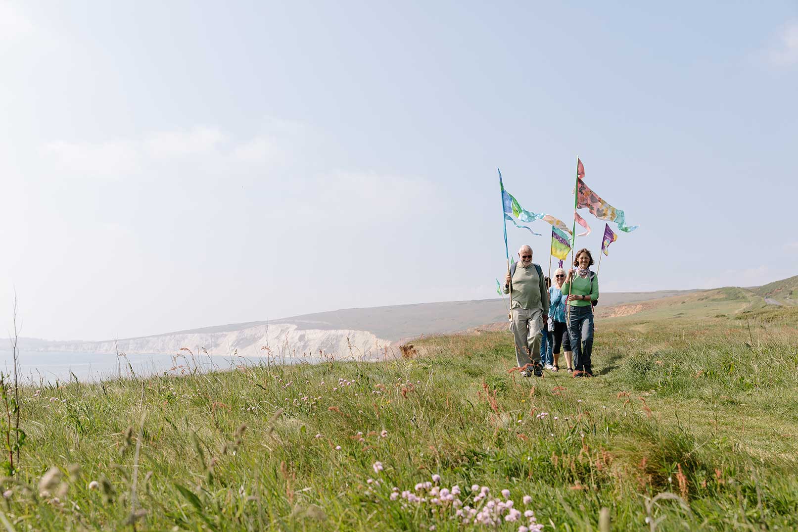 Small group of walkers walking along a coastal path carrying hand painted flags with bright green grass in the foreground, chalk cliffs in the background and a vast pale blue sky