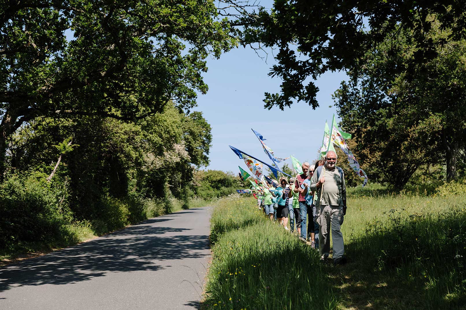 Walkers in procession carrying hand painted flags through rural countryside on grass verge with trees all around