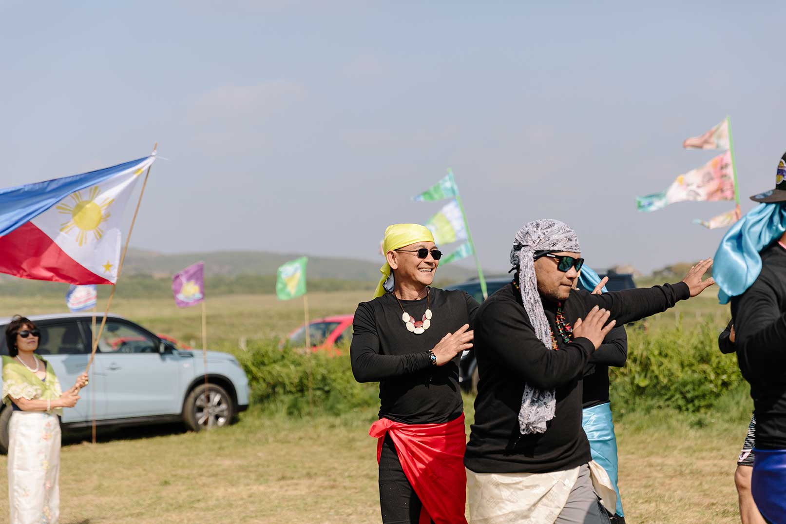 Men dancing a traditional Pilipino dance on a sunny day in a coastal field with painted flags in the background