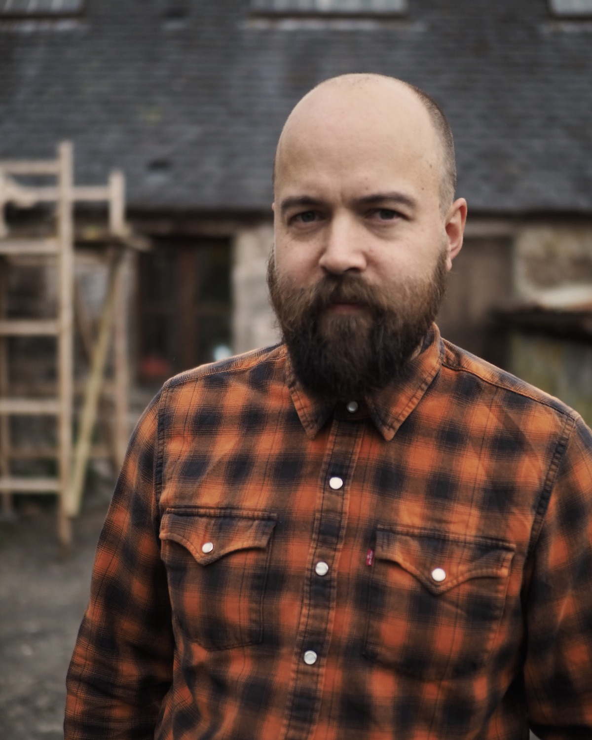 Photo of a male artist wearing an orange and black checked shirt outside in front of a barn