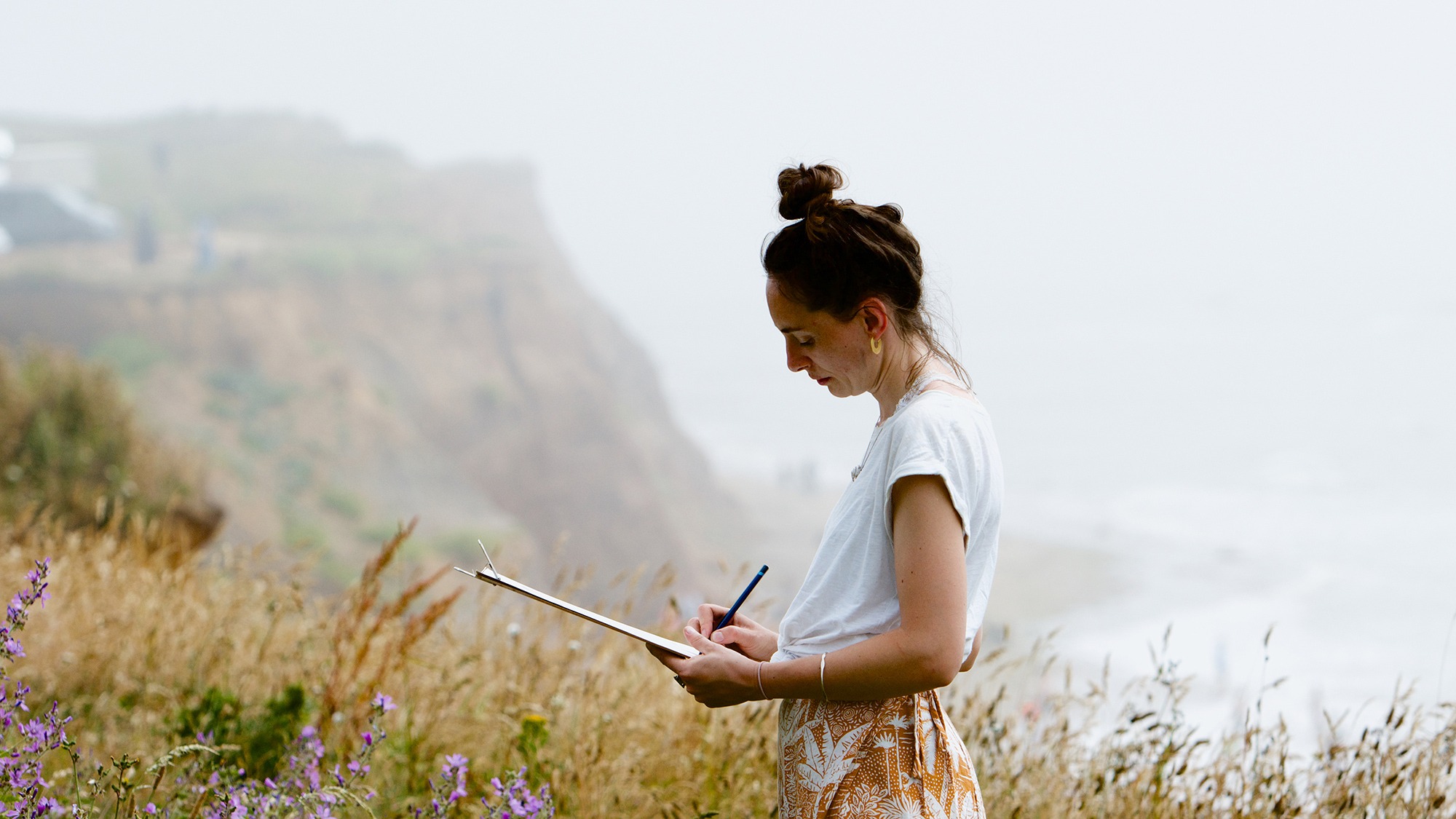 Photo of a female artist in summer clothes amongst coastal wildflowers with misty cliffs and sea in the background