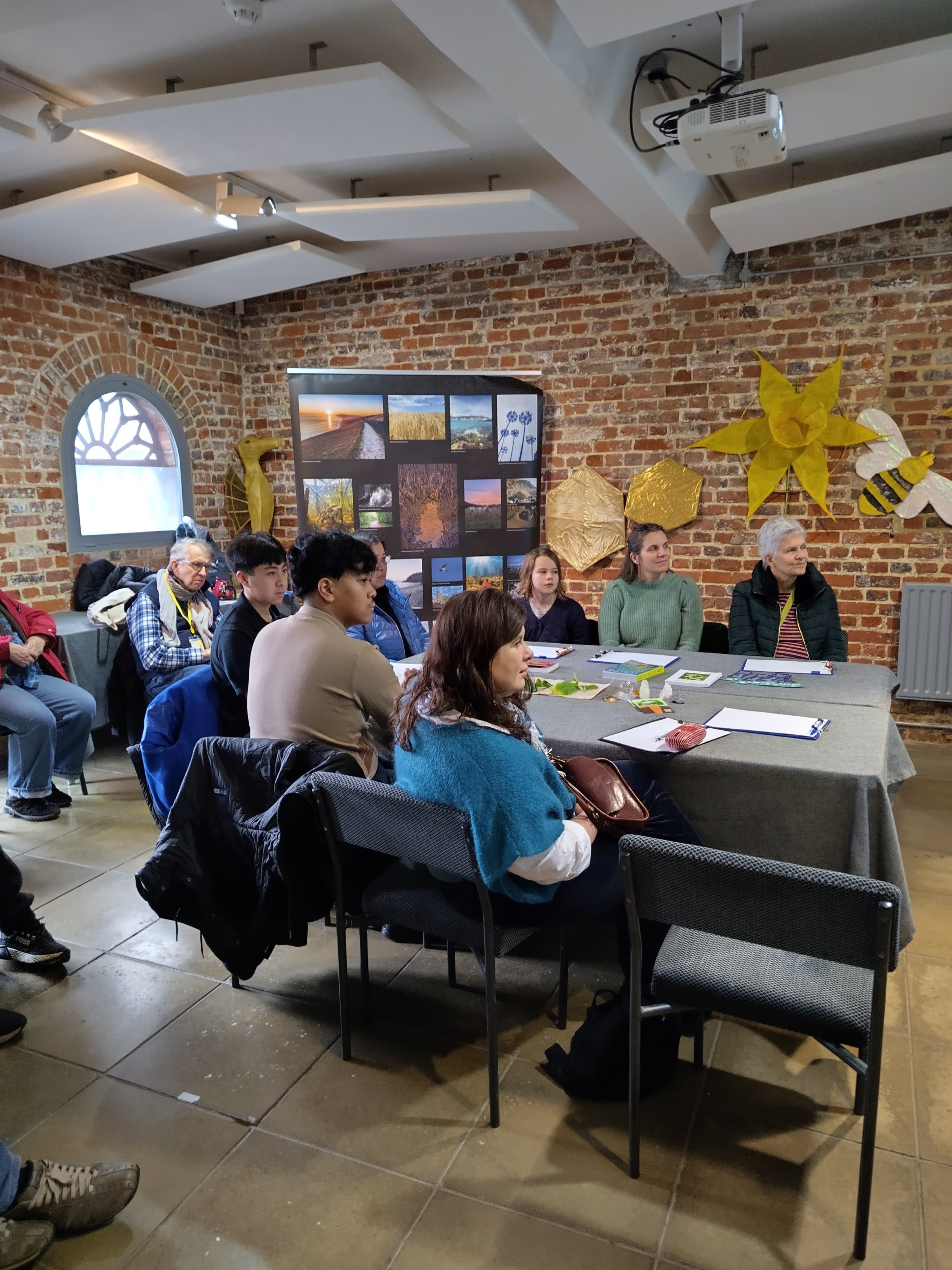 Workshop participants of all ages sat around a table listening to poetry. Nature images in the background in front of brick wall
