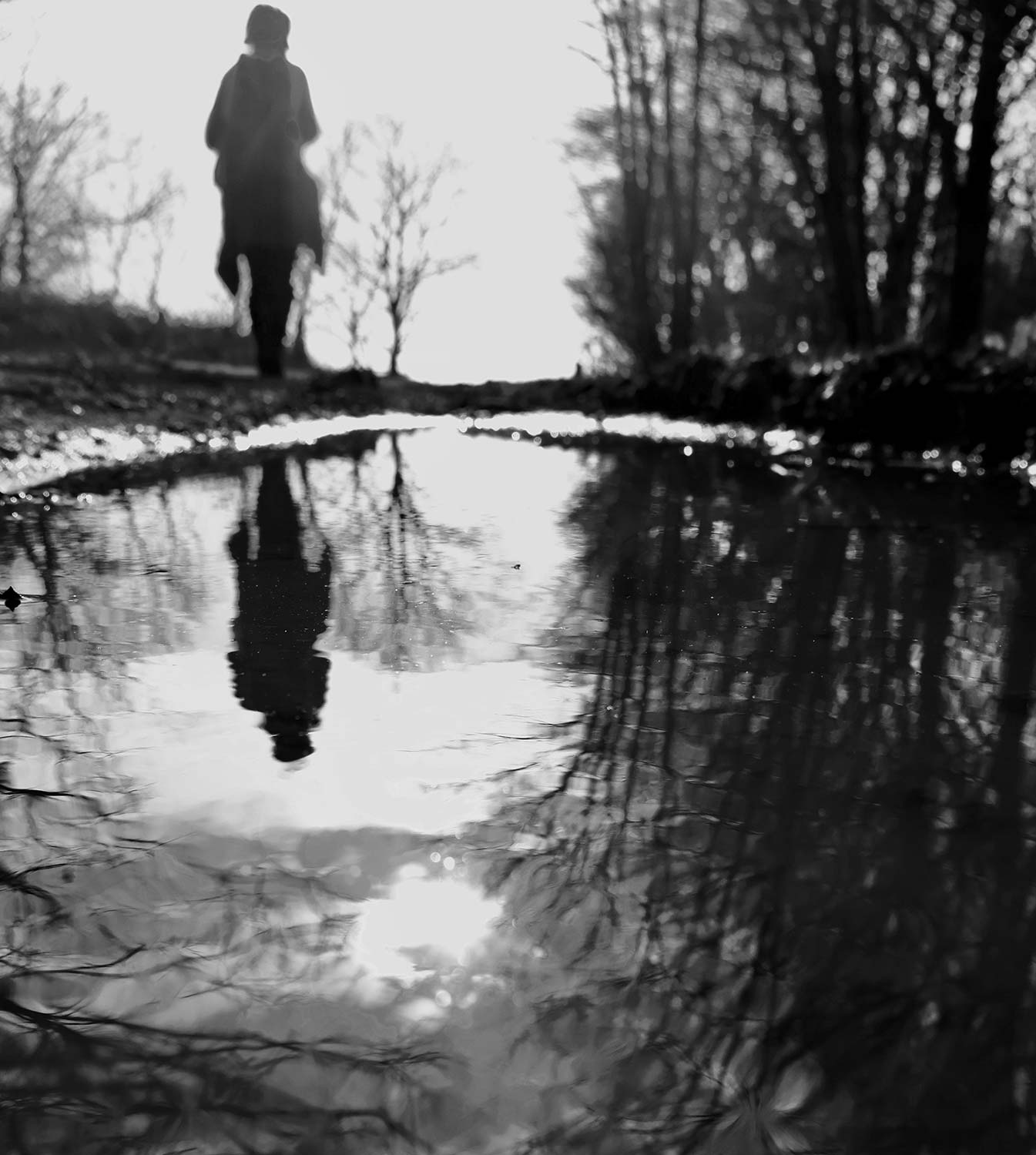 Black and white photo of a blurred figure in the distance reflected in a puddle in the foreground, surrounded by trees