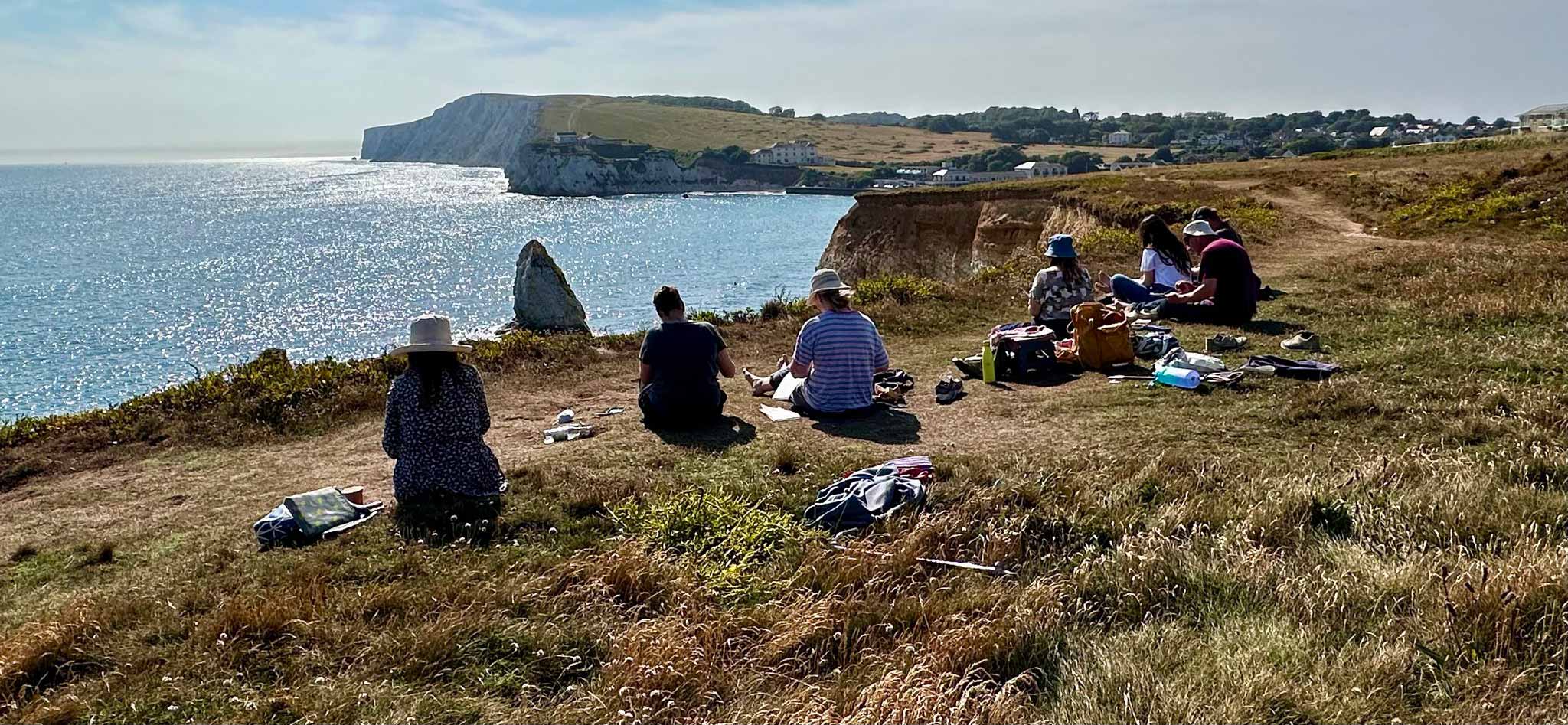 People sat on cliff top drawing with the sun sparkling on the water